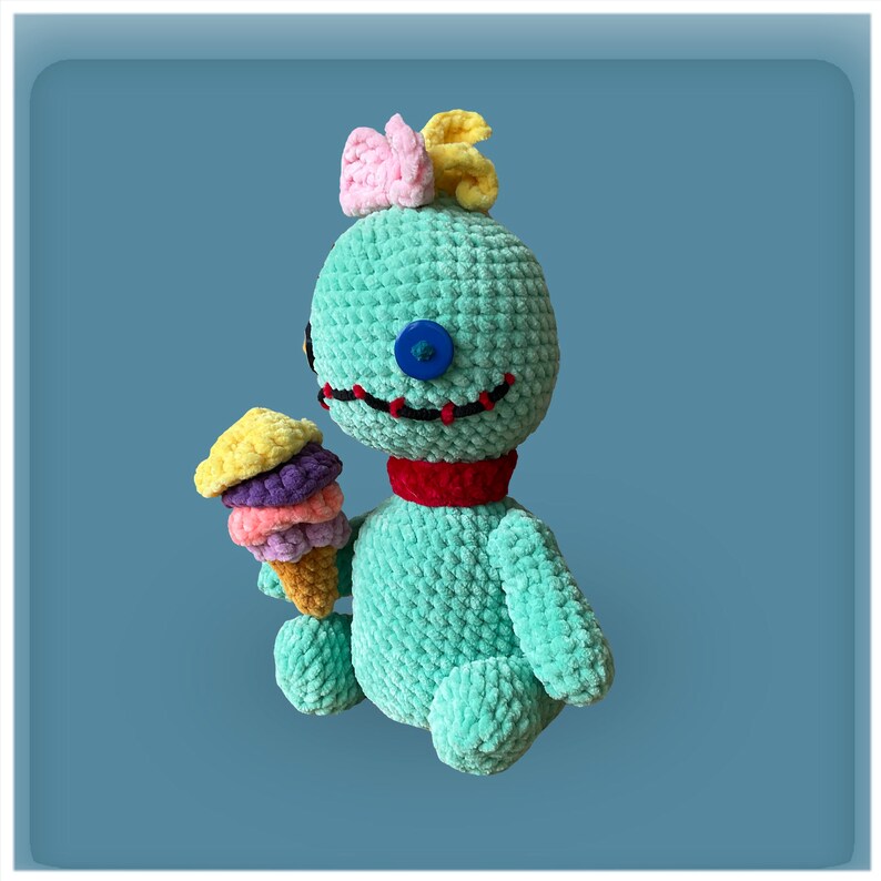 May include: A green crocheted plush toy with a red scarf and a single blue button eye. The toy is holding a multi-colored ice cream cone.