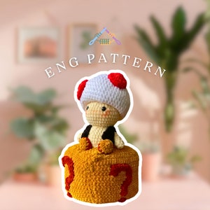 May include: Crocheted mushroom character sitting on a crocheted block with the number 37 on it. The mushroom character has a white body, red cap, and black eyes. The block is yellow with red numbers.