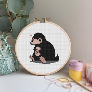 May include: A cross-stitch embroidery hoop with a white background and a black and pink Niffler design. The Niffler is sitting on its haunches with its paws crossed and a small pink heart above its head.