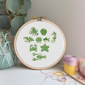 May include: A cross-stitch embroidery hoop with green sea creatures, such as an octopus and seahorse, on a white grid. The wooden hoop is displayed on a white surface. A vase and spools of thread are in the background.