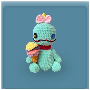 May include: A teal blue crocheted plush toy with a pink bow and red scarf, holding a multi-colored ice cream cone. The toy has black stitching around the eyes and mouth, and two button eyes.