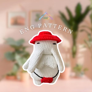 May include: White crocheted amigurumi toy with a red hat and a red belt. The toy has a long nose and large eyes. The toy is sitting on a table with a blurred background.