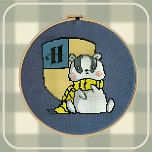 May include: A cross-stitch embroidery hoop with a white badger wearing a yellow scarf and a blue and gold crest with the letter "H" on it.