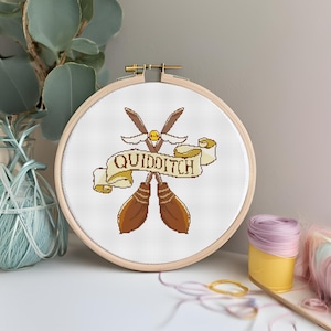 Peut inclure: Un cerceau de broderie blanc avec un motif brodé de deux balais dorés avec des ailes et une bannière qui dit "QUIDDITCH".