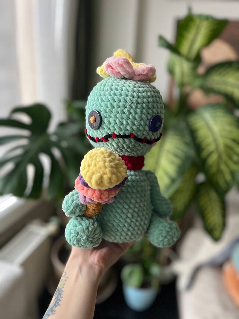May include: A green crocheted plush toy with a pink flower on its head, holding a multi-colored ice cream cone. The toy has black stitching around its eyes and mouth, and two buttons for eyes.