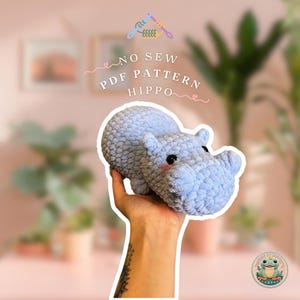 Puede incluir: Un juguete de hipopótamo de crochet azul claro. El juguete está siendo sostenido en una mano. El texto "NO SEW PDF PATTERN HIPPO" está escrito encima del juguete.
