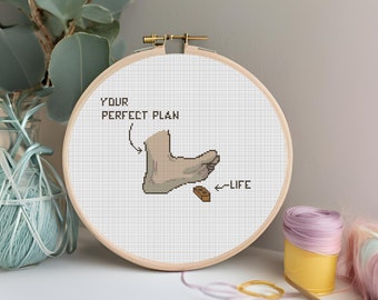 May include: A cross-stitch embroidery hoop with the text "YOUR PERFECT PLAN" and an image of a foot stepping on a brick labeled "LIFE". The design is in shades of beige and brown, set against a white background.