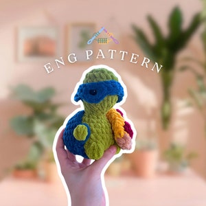 May include: A crocheted green turtle with a blue mask and a multi-colored shell. The turtle is being held by a hand. The image has the text "ENG PATTERN" above the turtle.