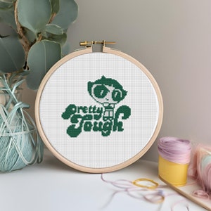 May include: A cross-stitch embroidery hoop featuring a green cartoon character and the words "Pretty Says Tough" stitched on white fabric. The wooden hoop is displayed on a white surface with craft supplies in the background.