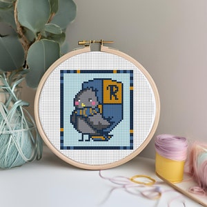 May include: A cross-stitch embroidery hoop with a pixelated image of a gray owl wearing a blue and gold hooded cloak with the letter "R" on the front. The owl is on a light blue background with a dark blue and gold border.