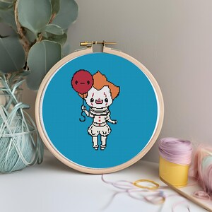 May include: A blue embroidery hoop with a pixelated image of a clown holding a red balloon. The clown is wearing a white and red costume with a black belt. The balloon has a frowning face.