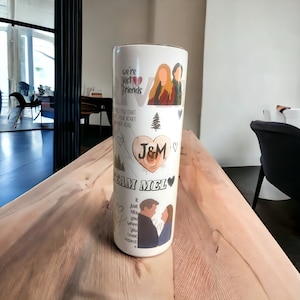 May include: A white cylindrical tumbler with a printed design. The design includes illustrations of people, hearts, and trees, along with the text "we're just friends", "J&M", and "Team Mel". The tumbler is displayed on a wooden surface.