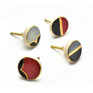 May include: Set of four ceramic cabinet knobs with a white base and black and gold geometric designs. Two knobs have a red accent, and two have a grey accent.