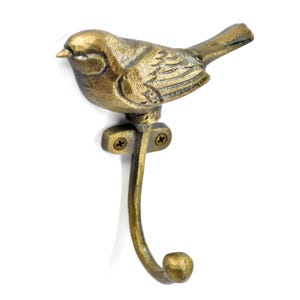 May include: A bronze-coloured metal wall hook shaped like a bird. The bird sits above a curved hook, attached to a mounting plate with two screws. This decorative hook is designed for hanging items, such as coats or bags.