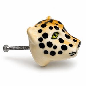 Peut inclure: Un bouton de meuble en céramique en forme de tête de léopard avec une base crème, des taches noires et un œil vert. Le bouton est doté d'une tige métallique argentée dépassant à l'arrière pour une fixation sur des meubles ou des placards.