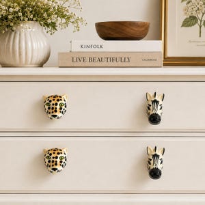May include: Four ceramic drawer knobs shaped like animal heads attached to a white dresser. Two knobs feature a leopard design with black spots and green eyes, while the other two knobs feature a black and white striped zebra design.