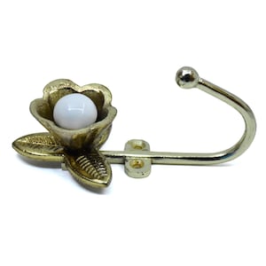 Könnte beinhalten: Ein dekorativer Wandhaken aus goldfarbenem Metall. Er zeigt eine Blume mit einem weißen, kugelförmigen Zentrum. Der Haken erstreckt sich von der Blume und hat ein abgerundetes Ende. Ideal zum Aufhängen von Gegenständen.