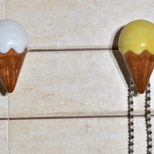May include: Two ceramic ice cream cone shaped hooks, one white and one yellow, mounted on a tiled wall. The yellow cone has a silver chain hanging from it.