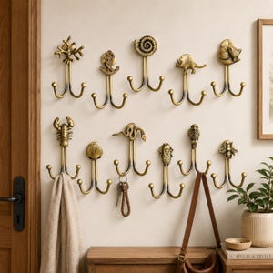 Puede incluir: Una colección de diez ganchos de pared en tono latón antiguo, cada uno con un adorno superior único que incluye una rama de coral, ancla, ammonite, dinosaurio, zorro, langosta, concha, serpiente, búho y abeja, montados en una pared clara.