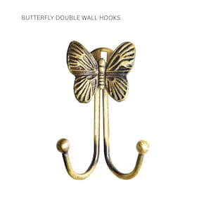 Puede incluir: Un gancho de pared decorativo con diseño de mariposa. El gancho es de metal con acabado bronce. La mariposa está centrada sobre dos ganchos curvos, cada uno con una pequeña bola en el extremo. El texto "BUTTERFLY DOUBLE WALL HOOKS" está en la parte superior.