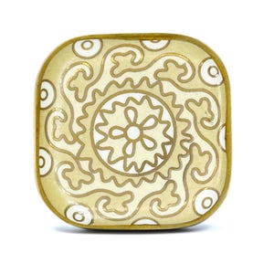 Puede incluir: Un plato cuadrado de cerámica con un diseño floral blanco y dorado. El plato tiene un borde elevado y un acabado liso y brillante.