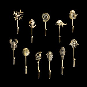 May include: A collection of decorative brass hooks featuring various designs, including a coral, an anchor, a nautilus shell, a dinosaur, a fish, a lobster, a seashell, a snake, a cicada, an owl, and a bee. Each hook has a single prong for hanging items.