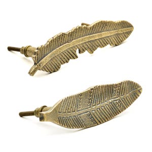 Puede incluir: Dos tiradores de armario en forma de pluma de color latón antiguo. Los tiradores tienen patrones detallados de vetas de plumas y roscas para la instalación. El herraje está sobre un fondo blanco.
