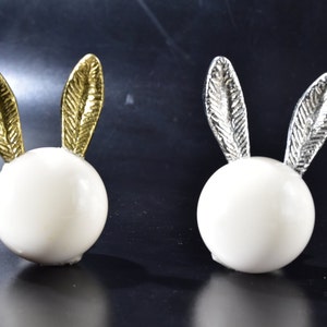 Puede incluir: Dos figuritas de conejo de cerámica blanca con orejas doradas y plateadas. El conejo de la izquierda tiene orejas doradas y el conejo de la derecha tiene orejas plateadas.