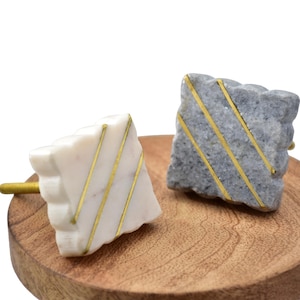 Puede incluir: Dos tiradores decorativos para armarios. Uno es blanco con rayas doradas, el otro es gris con rayas doradas. Ambos tienen forma cuadrada con una superficie texturizada. Los tiradores se muestran sobre una superficie de madera.
