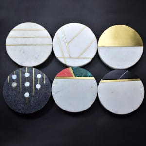 May include: Six circular coasters with decorative designs. The coasters feature white marble, dark grey stone, and gold accents. Some have geometric patterns, while others have inlaid stone and metal details. The coasters are arranged on a black surface.