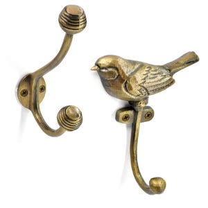 Op de afbeelding: Twee antieke messing wandhaken. Eén haak heeft een spiraalvormig ontwerp aan de boven- en onderkant. De andere haak is in de vorm van een vogel. Beide haken hebben een gebogen ontwerp en zijn met schroeven bevestigd.