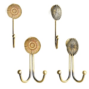 Pode incluir: Quatro ganchos de parede em latão antigo. Dois ganchos simples e dois duplos. Cada gancho apresenta um design floral decorativo no topo. Os ganchos são montados em um fundo branco.
