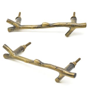 Può includere: Due pomelli per mobili color bronzo a forma di rami d'albero. Ogni pomello ha una superficie strutturata e una vite per il montaggio. I pomelli sono su uno sfondo bianco.