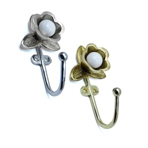 Puede incluir: Dos ganchos de pared decorativos de metal. Uno es plateado, el otro dorado. Cada gancho presenta un diseño floral con un centro esférico blanco. Los ganchos están diseñados para colgar objetos y añaden un toque de elegancia.