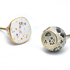 Op de afbeelding: Twee keramische kastknoppen. Eén is vierkant met een witte achtergrond en kleurrijke stippen. De andere is rond met een witte achtergrond en een zwart bloemenontwerp.
