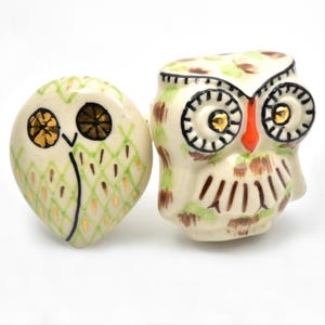 May include: Two ceramic owl figurines. One is oval-shaped with a green and gold diamond pattern. The other is upright with a white, green, and brown pattern. Both have black and gold eyes and are on a white background.