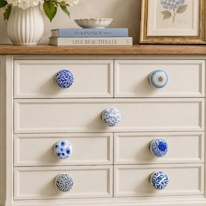 May include: A cream-colored wooden dresser featuring seven drawers, each equipped with a unique round ceramic knob. The knobs display various blue and white patterns, including floral, leaf, and geometric designs, providing a decorative accent.