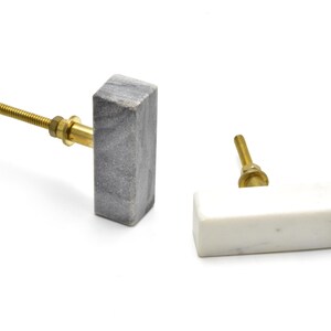 May include: Two rectangular cabinet knobs made of marble. One is grey with a brass screw and the other is white with a brass screw.