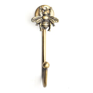 May include: A brass-toned wall hook with a bee motif. The hook has a long stem with a rounded end and a curved hook. The bee is at the top, adding a decorative element. The hook is suitable for hanging items and measures approximately 15 cm in length.