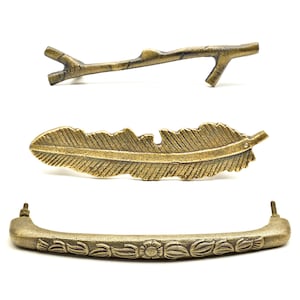 May include: Three bronze-coloured cabinet pulls. The top pull is shaped like a twig with two branches. The middle pull is a feather shape. The bottom pull is a curved handle with floral and leaf detailing.