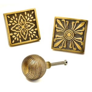 以下が含まれることがあります： アンティーク真鍮製のキャビネット金具3点セット。2つの正方形のノブは、花柄とサンバーストのデザインが特徴です。リブ加工が施された丸いノブも含まれています。すべてのパーツはヴィンテージ感があります。
