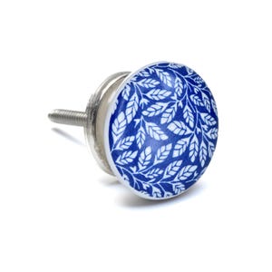 Può includere: Un pomello rotondo per mobili con un motivo a foglie blu e bianco sulla superficie. Il pomello presenta una faccia circolare liscia e una base in metallo color argento con una vite filettata che sporge dal retro per l'installazione.