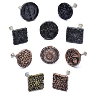 May include: A collection of decorative cabinet knobs in various shapes and designs. The knobs are in black, bronze, and gold tones, featuring floral, geometric, and textured patterns. Each knob has a metal screw for easy installation.