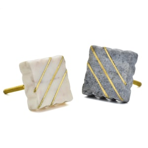 Könnte beinhalten: Zwei quadratische Schrankknöpfe. Einer ist aus weißem Marmor mit goldenen Streifen, der andere aus grauem Marmor mit goldenen Streifen. Beide haben goldfarbene Stiele.