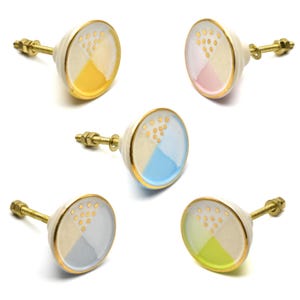 Może przedstawiać: Pięć ceramicznych gałek do szafek z złotymi akcentami. Każda gałka ma inny kolor: żółty, różowy, niebieski, jasnoniebieski i zielony. Gałki mają białe tło z złotymi kropkami i złotą obwódką.