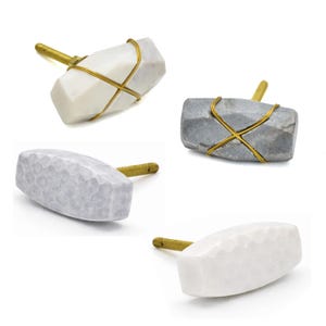 Puede incluir: Un conjunto de cuatro tiradores para armarios. Los tiradores presentan un aspecto similar a la piedra en blanco, gris y blanquecino. Cada tirador tiene un vástago de color latón y está envuelto con alambre dorado. Diseñados para muebles.