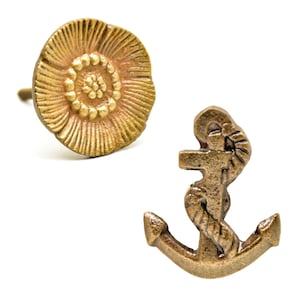 May include: Two decorative cabinet knobs. One is a flower shape with a detailed petal design and a brass finish. The other is an anchor shape with a rope detail and a brass finish. Both knobs are on a white background.