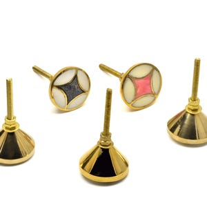 May include: Five decorative cabinet knobs. Two knobs have a circular design with a gold border and a central geometric pattern in pink and dark grey. The other three knobs have a gold conical base.