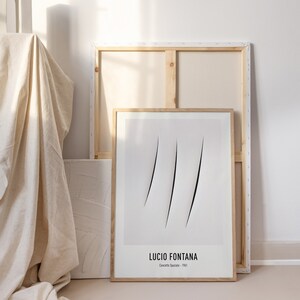May include: A framed black and white minimalist art print with a simple line design. The print is titled "Concetto Spaziale - 1961" by Lucio Fontana.