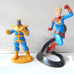 May include: Two action figures of Marvel characters. One figure is Thanos, a purple-skinned character in a blue and gold suit. The other is Captain Marvel, in a blue, red, and gold suit, posed mid-flight on a black stand.